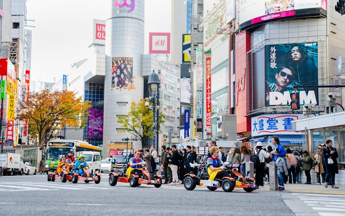 Tourists go-karting in Shibuya, Tokyo, surrounded by city buildings and billboards.