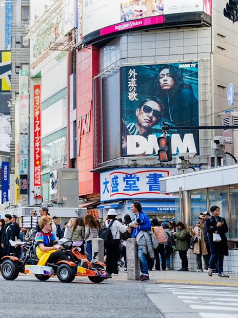 Tourists go-karting in Shibuya, Tokyo, surrounded by city buildings and billboards.