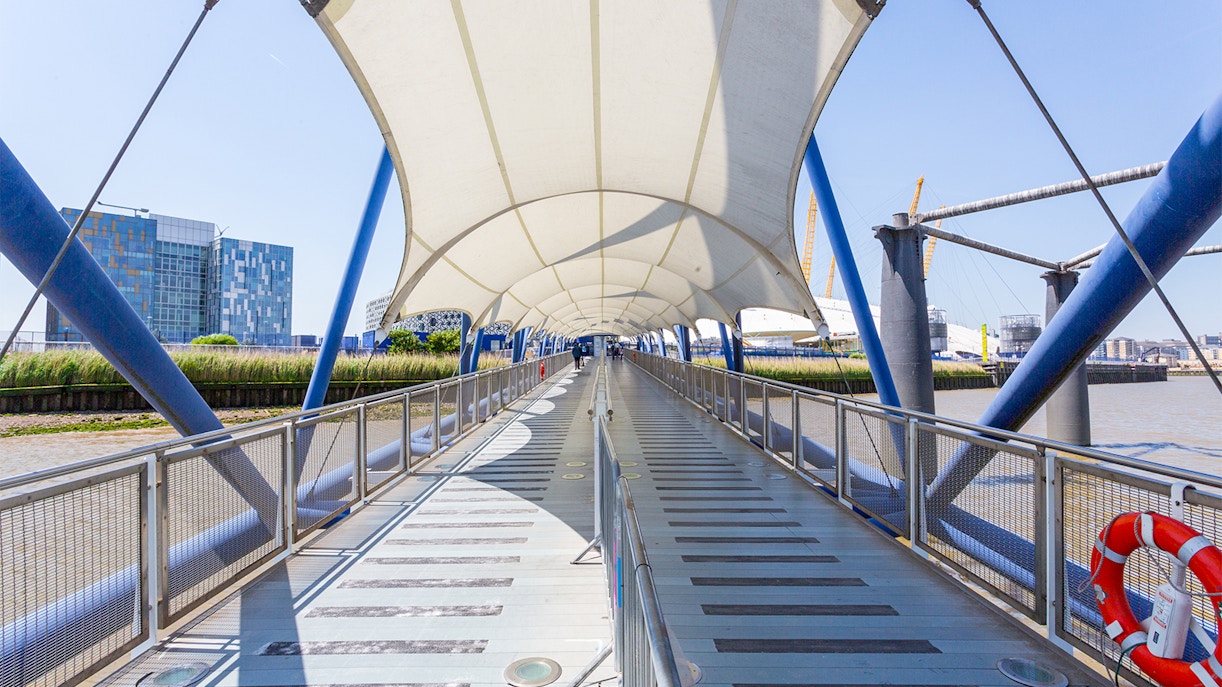 Covered walkway at Greenwich Pier with view of the Thames and modern buildings.