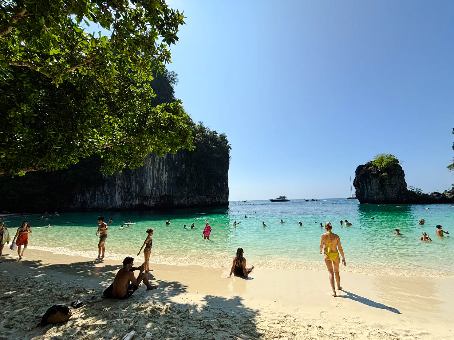 Tourists swimming and relaxing on a sandy beach at Hong Islands, Krabi, with boats in the distance.