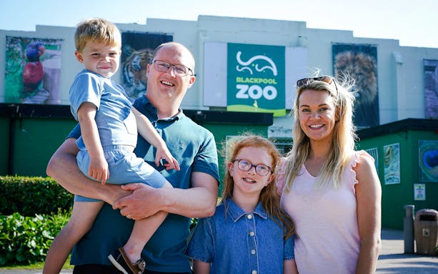 Family visiting Blackpool Zoo entrance with animal posters in background.