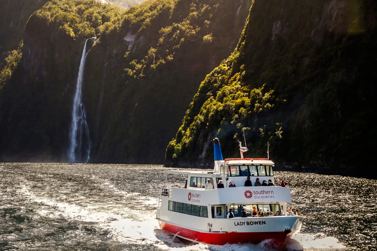 Cruise boat on Milford Sound with waterfall and lush cliffs, Queenstown tour.