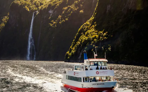 Cruise boat on Milford Sound with waterfall and lush cliffs, Queenstown tour.