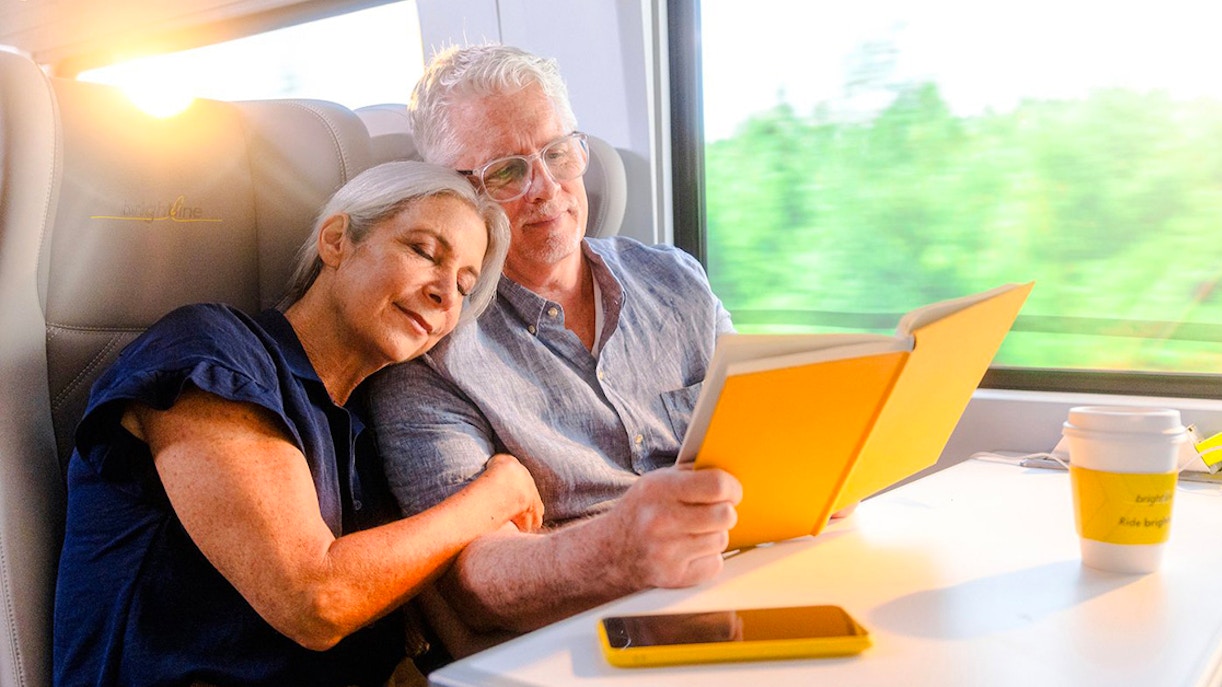 Couple relaxing on Brightline train from Miami to Orlando, reading and enjoying the journey.