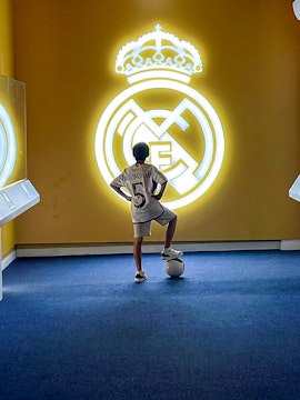 Child in a Real Madrid jersey stands with a soccer ball in front of the Real Madrid emblem at Real Madrid Park.