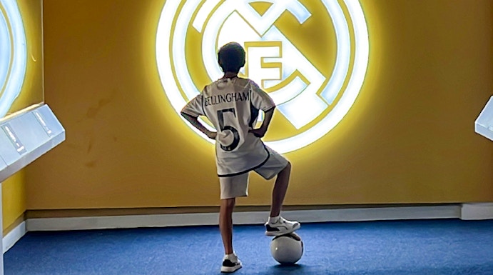 Child in a Real Madrid jersey stands with a soccer ball in front of the Real Madrid emblem at Real Madrid Park.