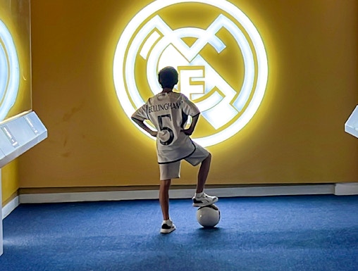 Child in a Real Madrid jersey stands with a soccer ball in front of the Real Madrid emblem at Real Madrid Park.