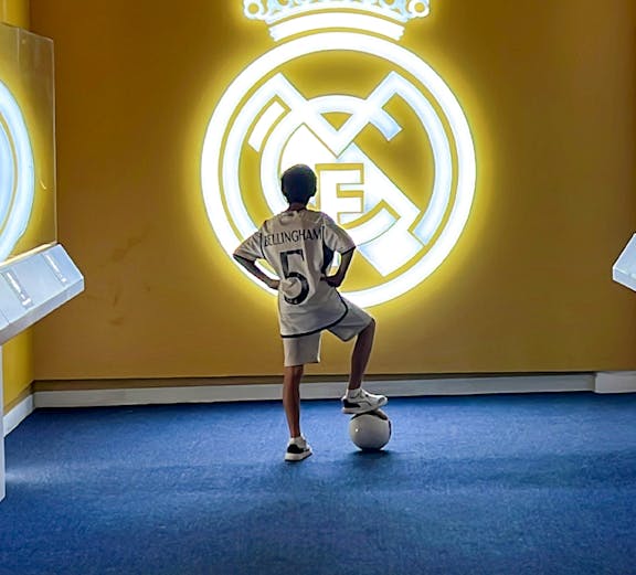 Child in a Real Madrid jersey stands with a soccer ball in front of the Real Madrid emblem at Real Madrid Park.