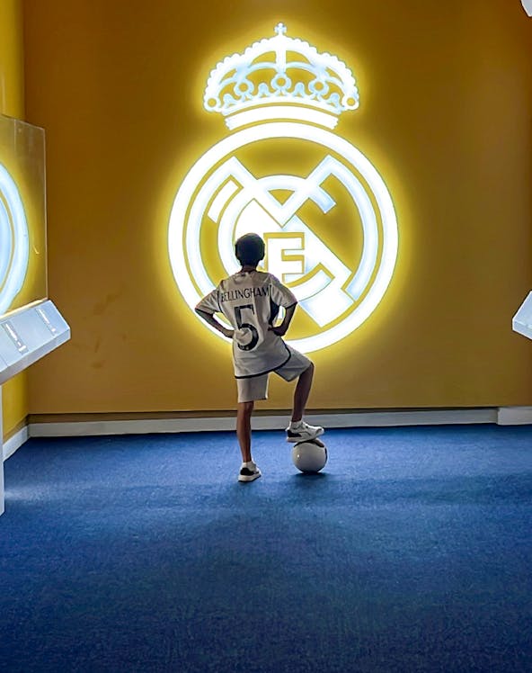 Child in a Real Madrid jersey stands with a soccer ball in front of the Real Madrid emblem at Real Madrid Park.