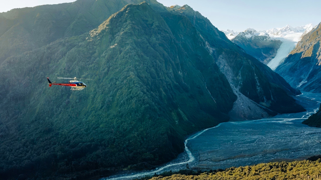 Helicopter flying over Franz Josef Glacier in New Zealand with lush mountains.