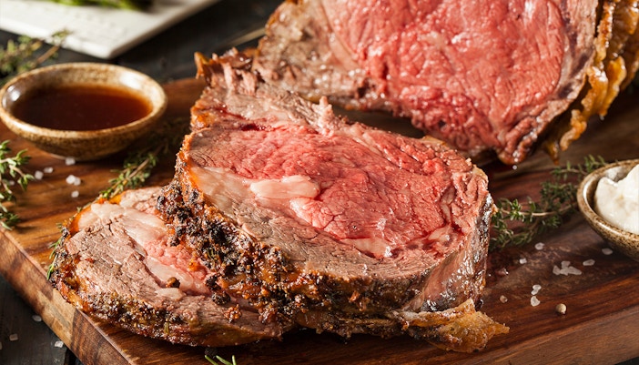 Roast beef slices on a wooden board with herbs and dipping sauce.