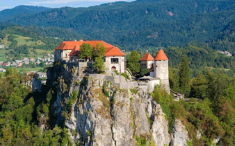 Bled Castle perched on a cliff with lush forest and mountains in Slovenia.