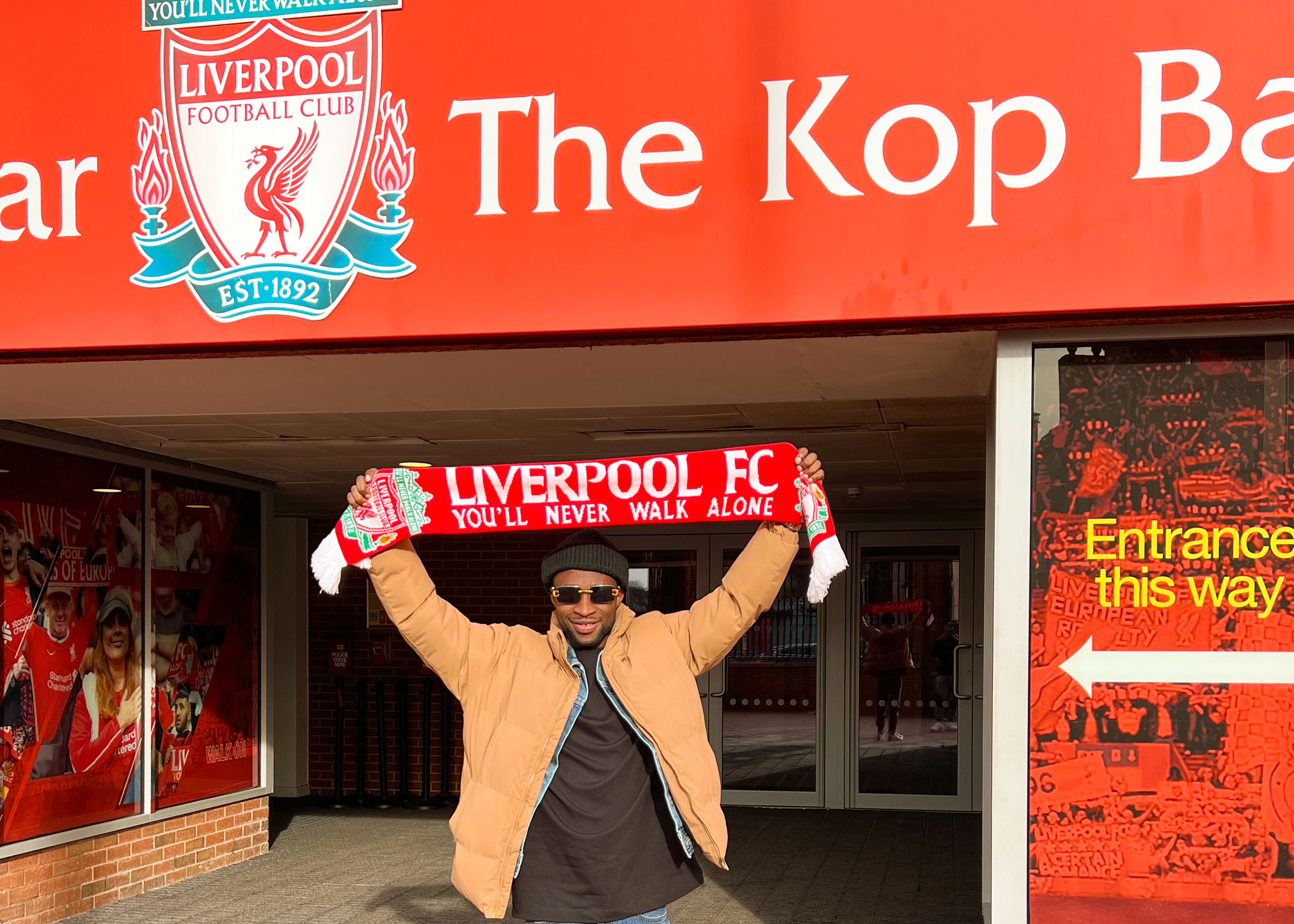 Person holding Liverpool FC scarf outside Anfield's Kop entrance.