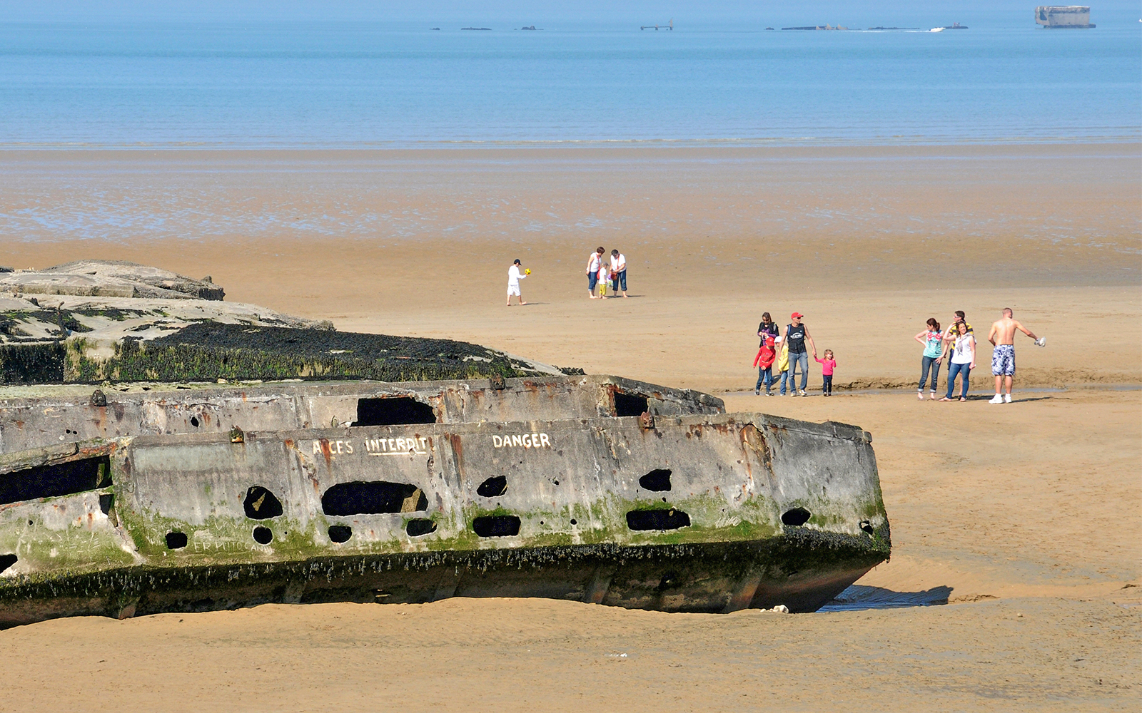 Normandy beach with WWII landing craft remains and tourists exploring the site.