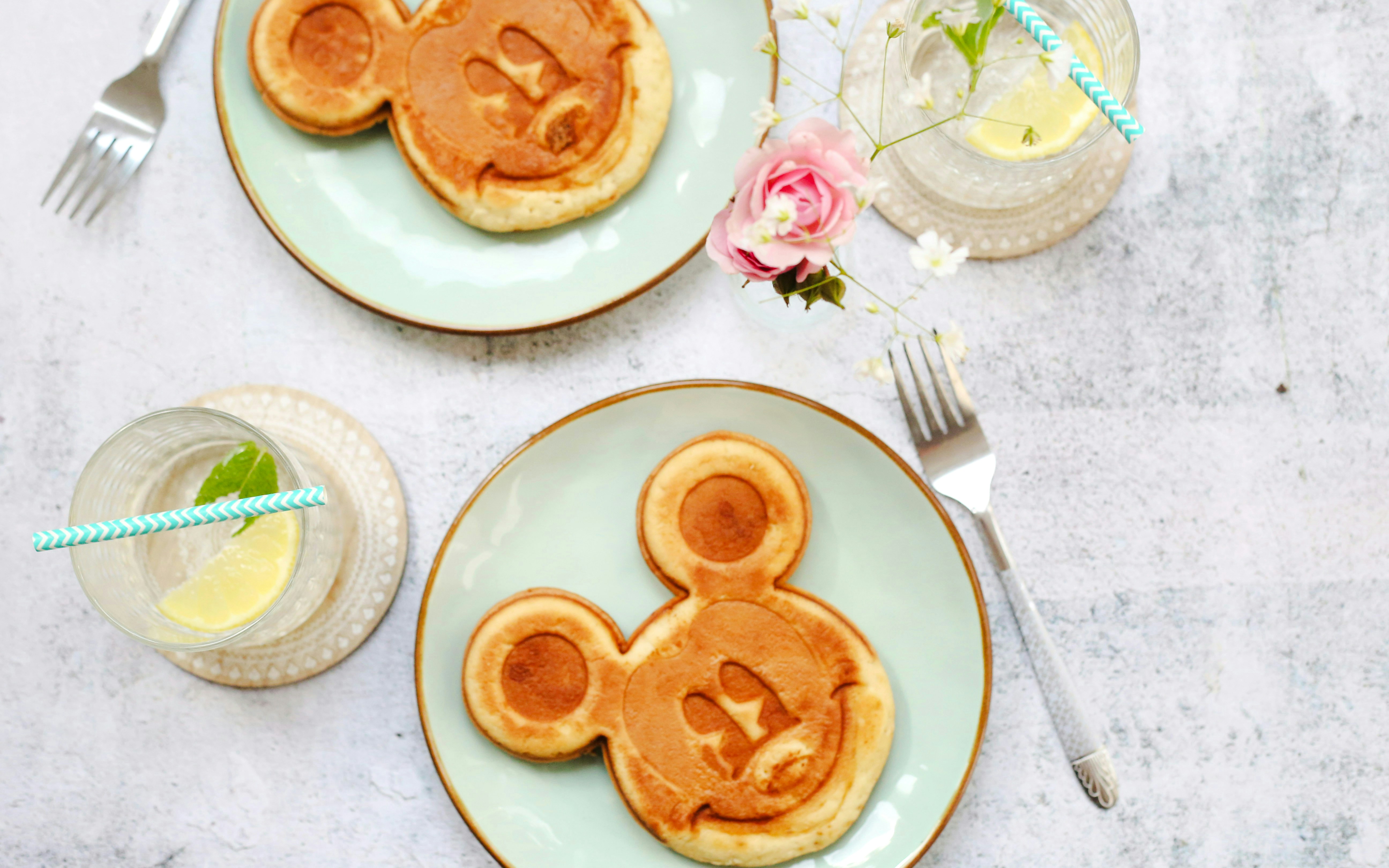 Mickey Mouse-shaped pancakes on plates with drinks and flowers.