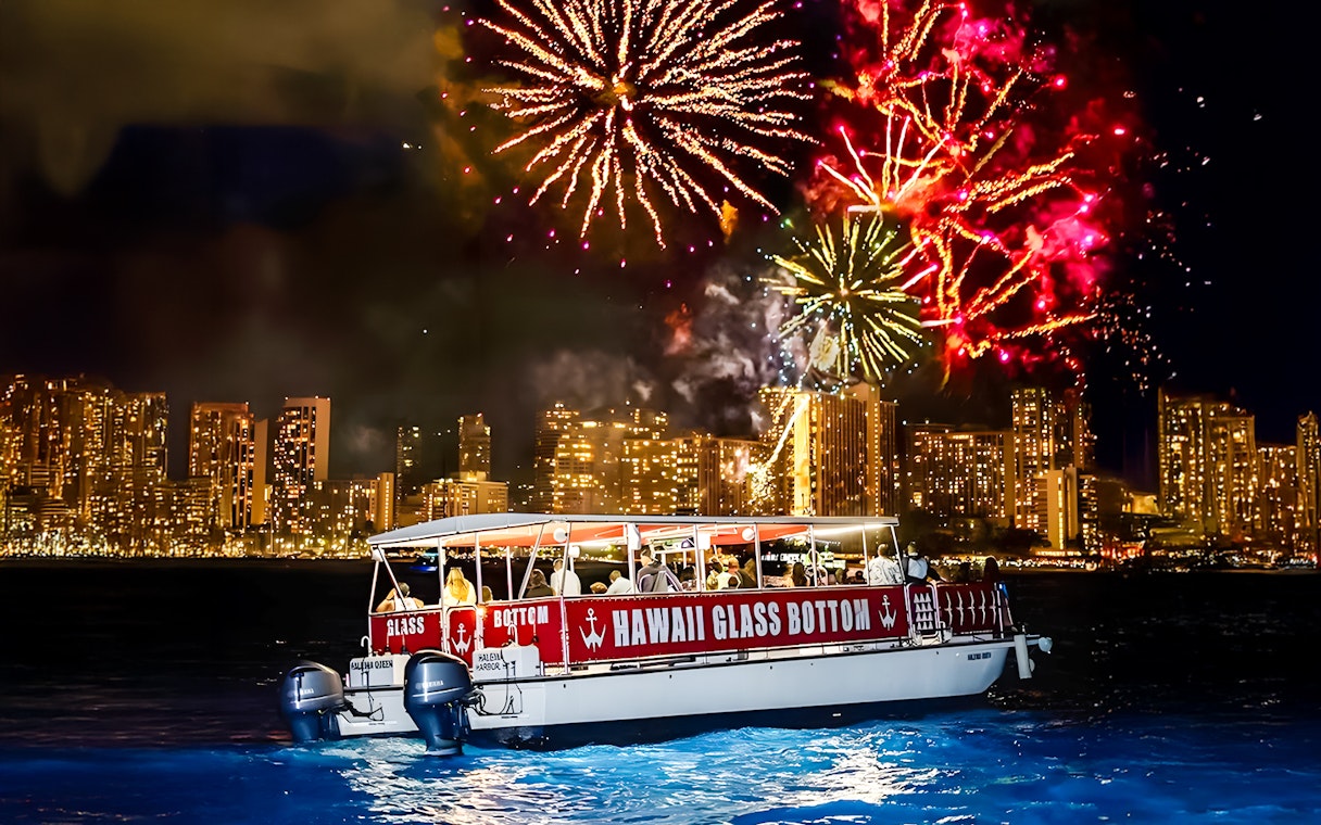 Glass-bottom boat "HAWAII GLASS BOTTOM" on water at night with fireworks over city skyline.