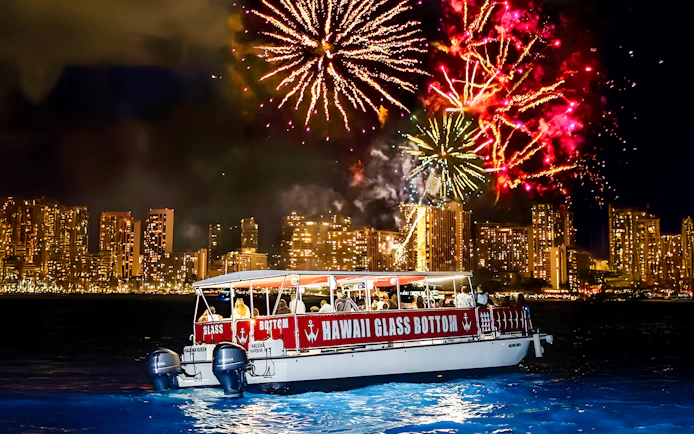 Glass-bottom boat "HAWAII GLASS BOTTOM" on water at night with fireworks over city skyline.