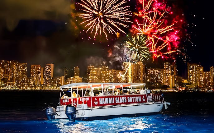 Glass-bottom boat "HAWAII GLASS BOTTOM" on water at night with fireworks over city skyline.