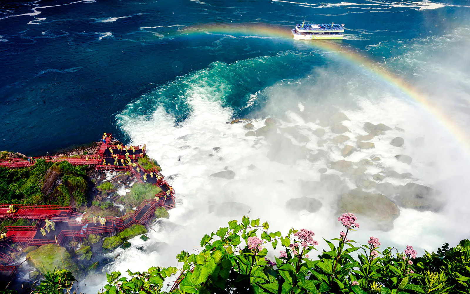 Visitors on red walkways near Niagara Falls USA with a rainbow and tour boat in the background.