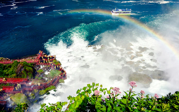 Visitors on red walkways near Niagara Falls USA with a rainbow and tour boat in the background.