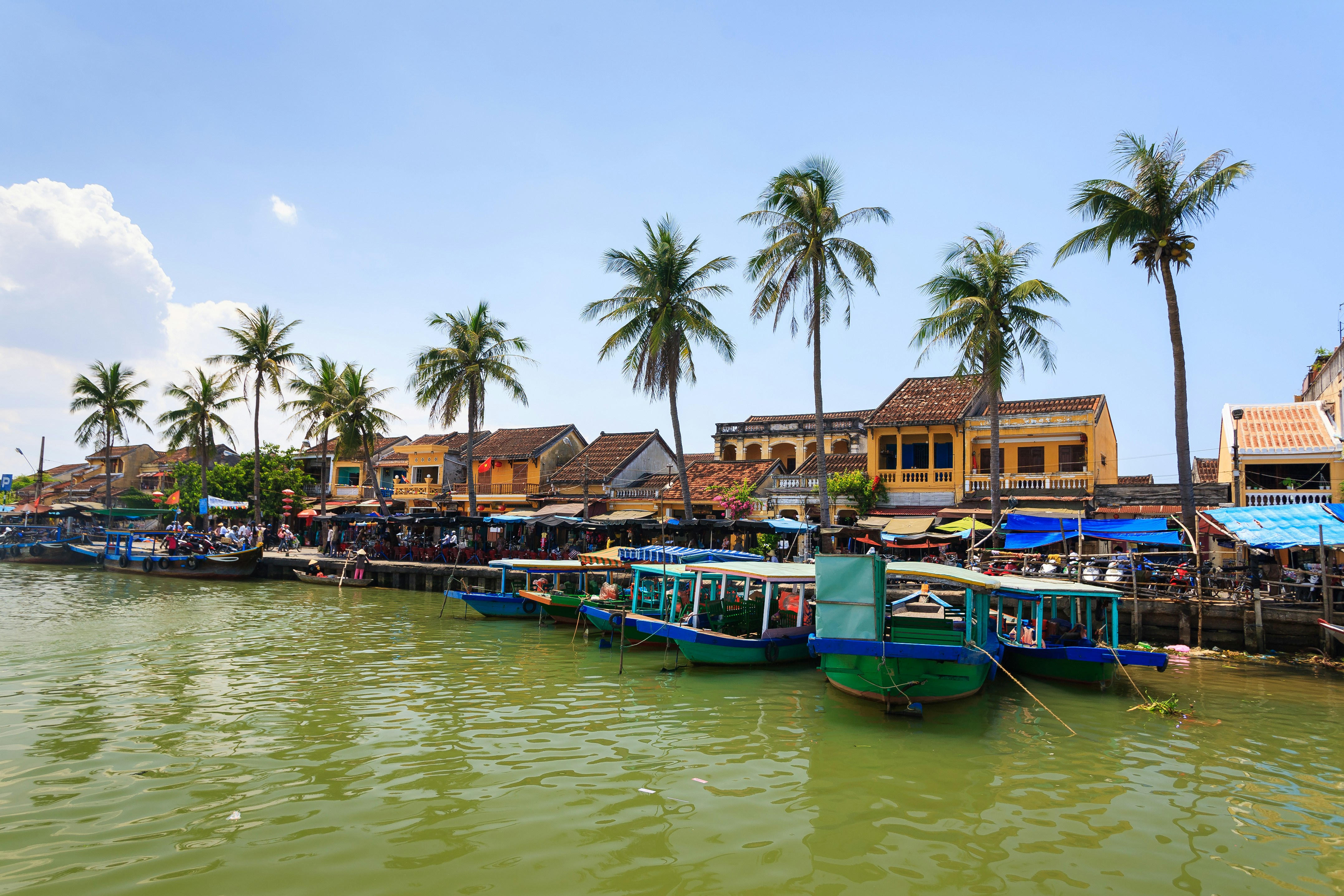 Boats docked at Bach Dang Wharf, Hoi An Ancient Town, with yellow buildings and palm trees.