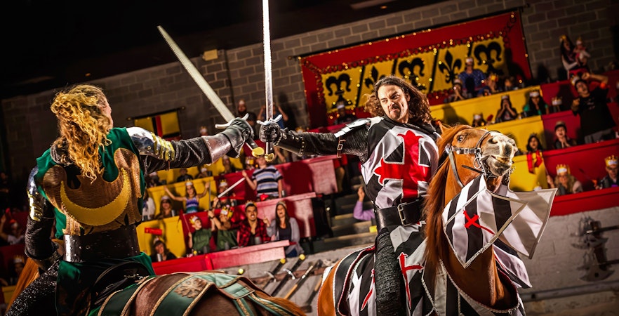 Knights jousting on horseback at Medieval Times Dinner and Show, with an audience in the background.