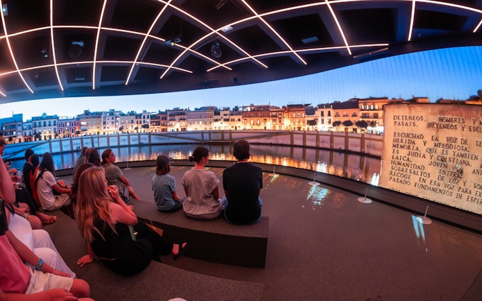 Visitors seated inside Setas de Sevilla, viewing a nighttime cityscape projection, Spain.
