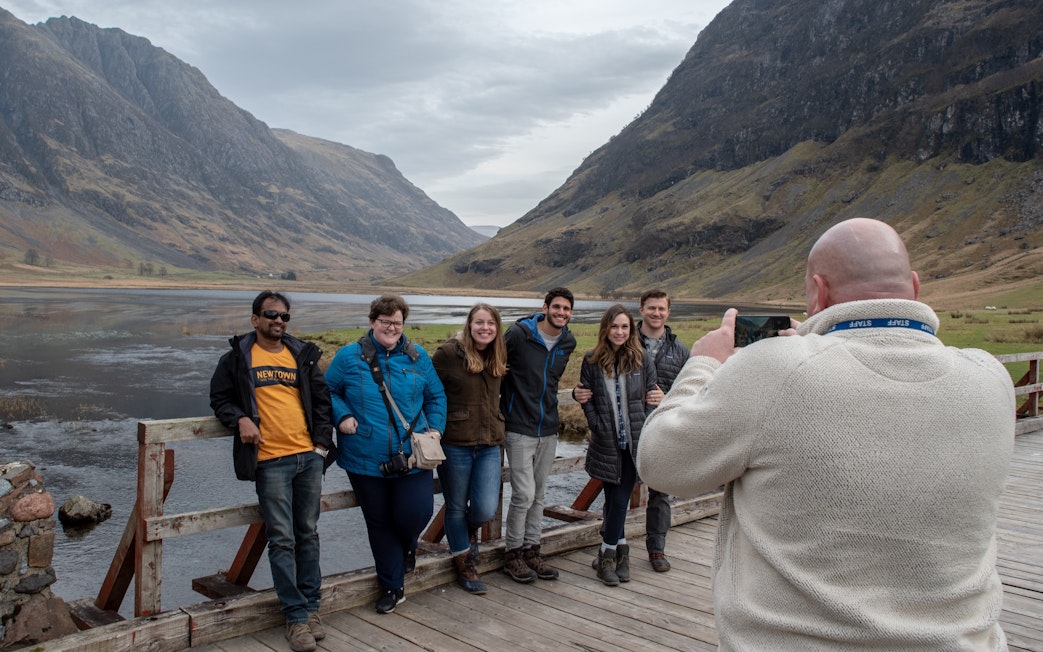 Group posing for a photo on a wooden bridge in Glencoe, Scotland, with mountains in the background.