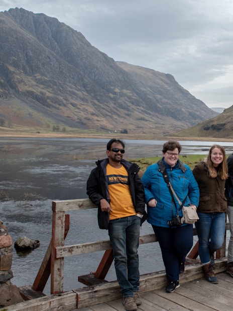 Group posing for a photo on a wooden bridge in Glencoe, Scotland, with mountains in the background.