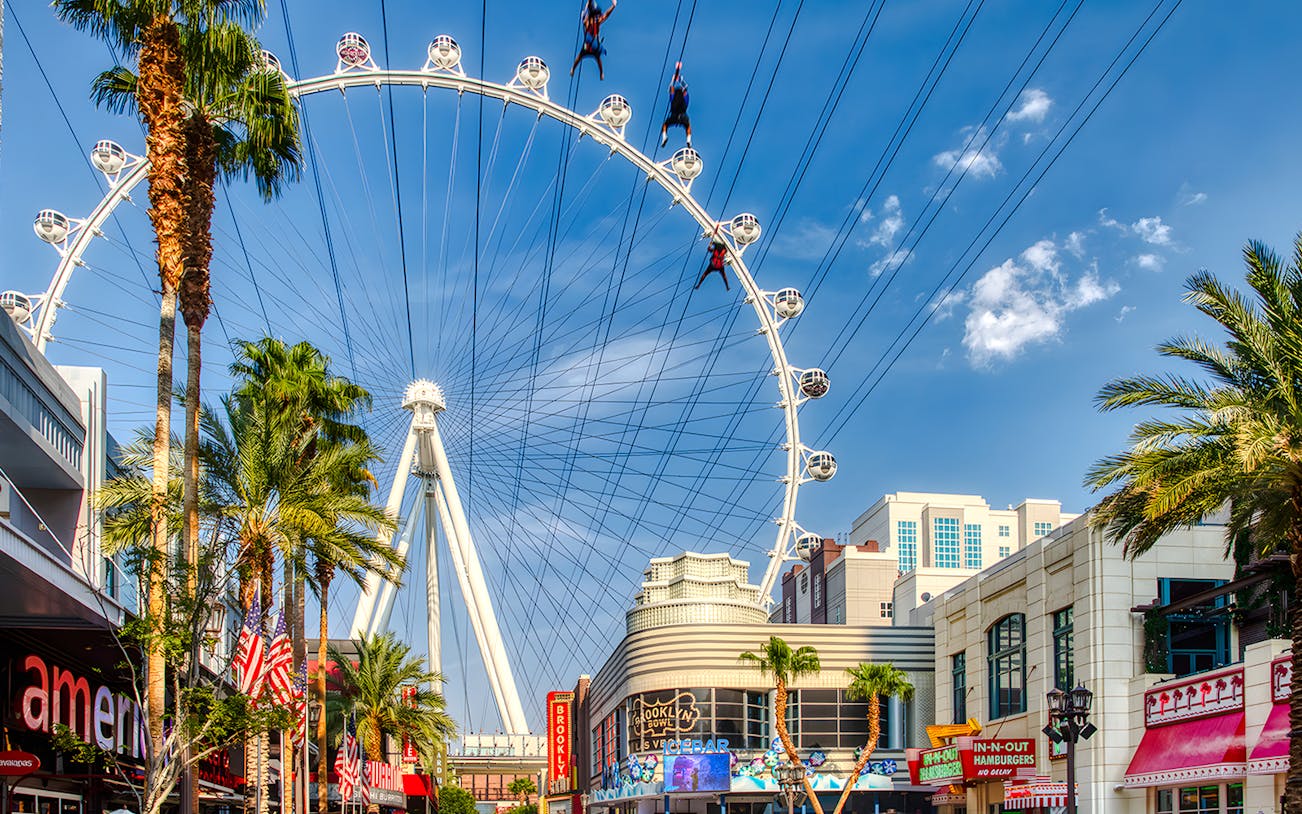 Fly LINQ Zipline riders above Las Vegas Strip with High Roller Ferris wheel in background.