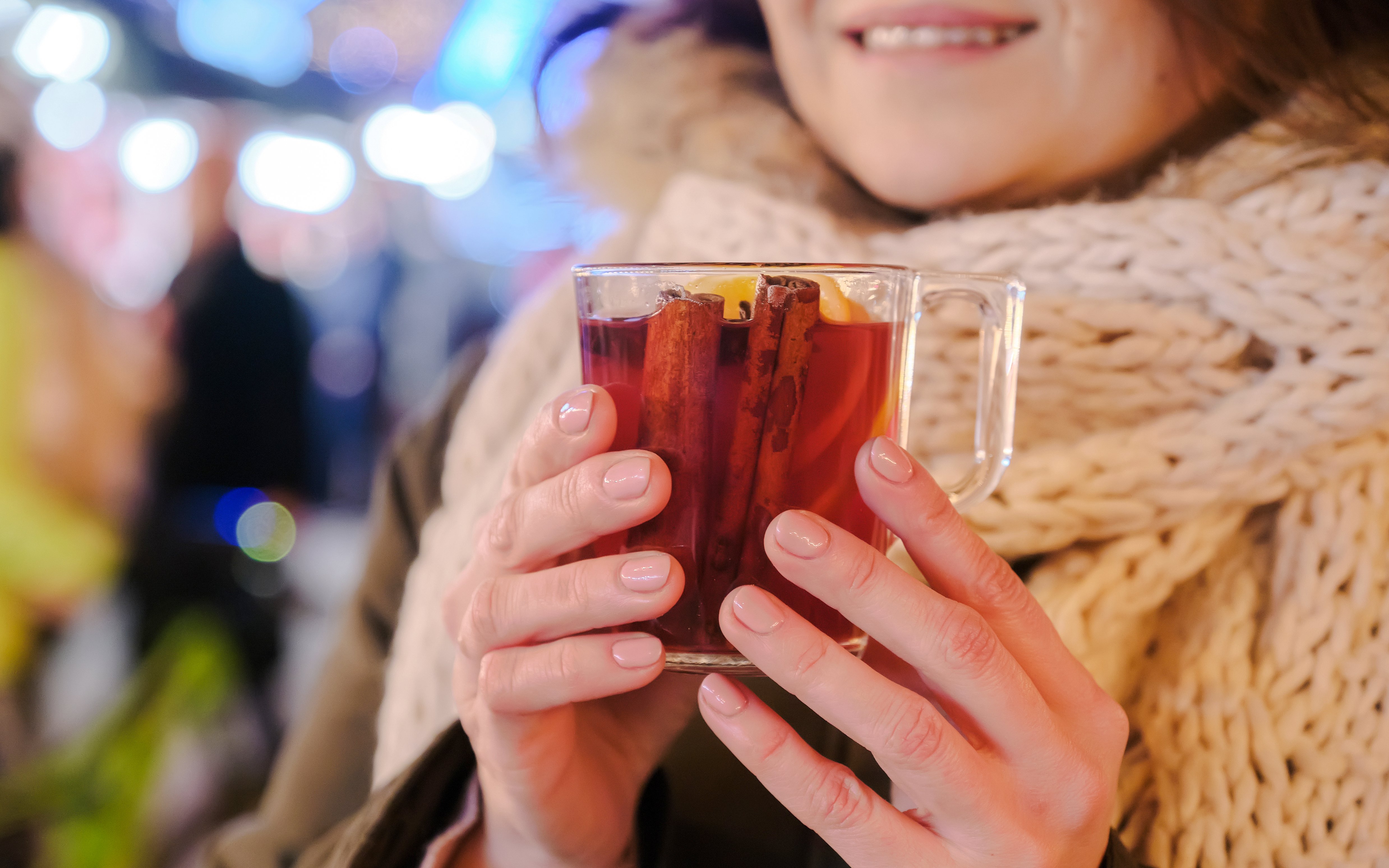 Mulled wine in clear mug with cinnamon sticks and lemon slice, held by woman in scarf.