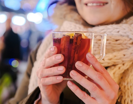 Mulled wine in clear mug with cinnamon sticks and lemon slice, held by woman in scarf.