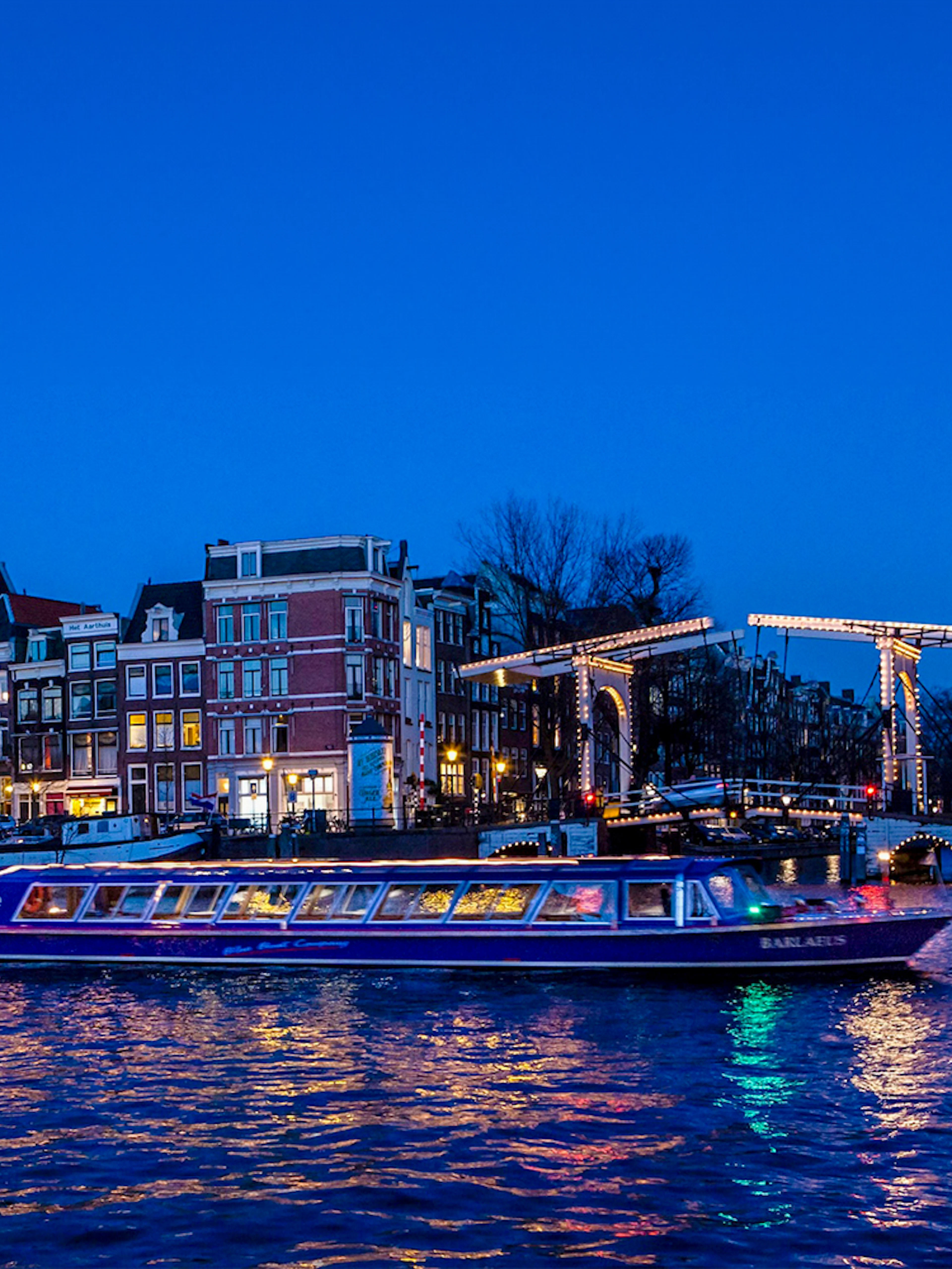 Amsterdam canal cruise boat passing under illuminated bridge at dusk.