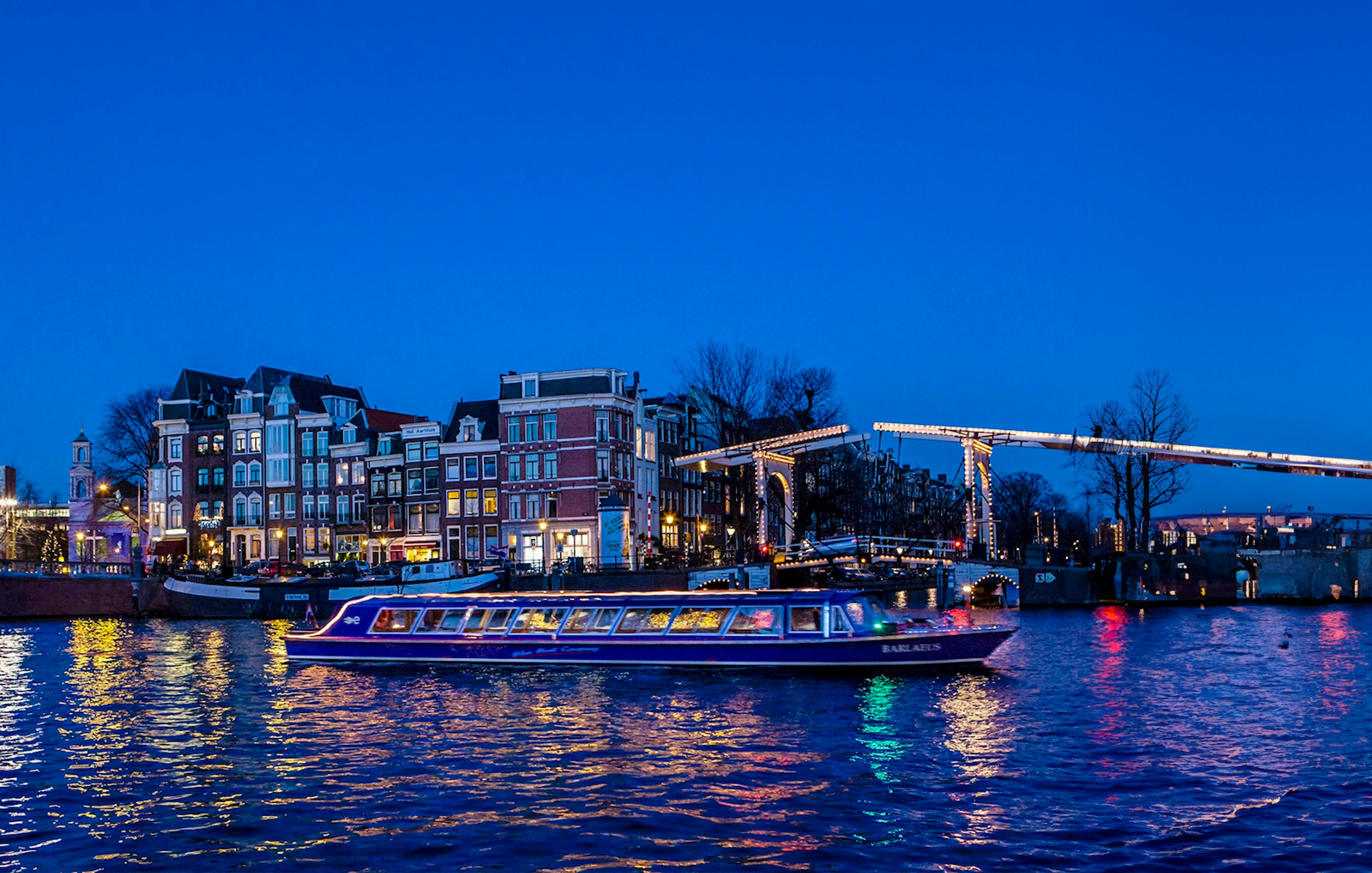 Amsterdam canal cruise boat passing under illuminated bridge at dusk.