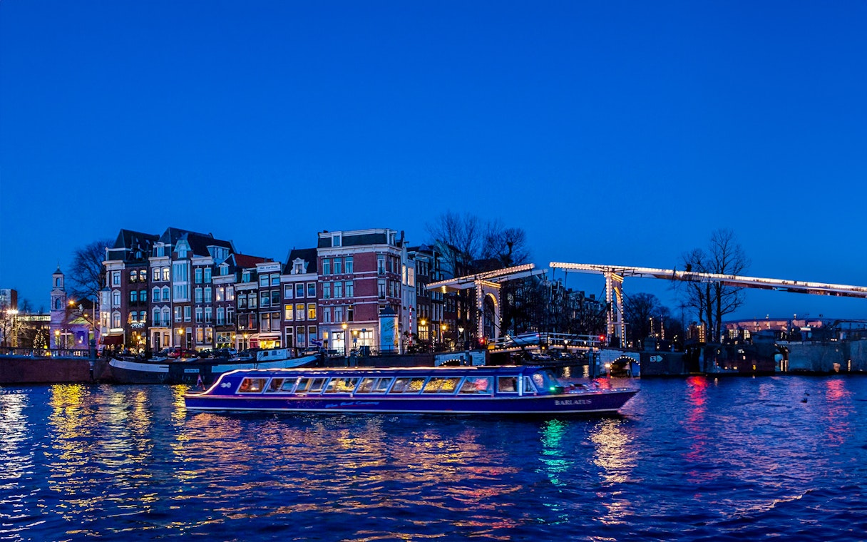 Amsterdam canal cruise boat passing under illuminated bridge at dusk.