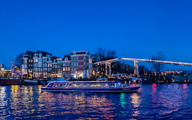 Amsterdam canal cruise boat passing under illuminated bridge at dusk.
