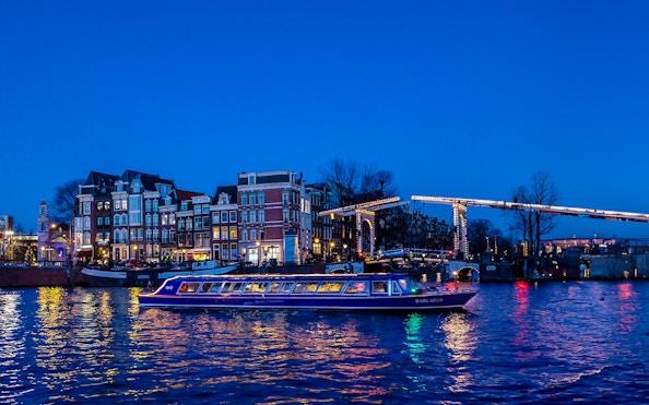 Amsterdam canal cruise boat passing under illuminated bridge at dusk.