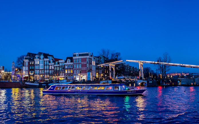 Amsterdam canal cruise boat passing under illuminated bridge at dusk.
