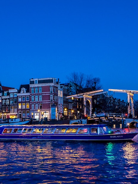 Amsterdam canal cruise boat passing under illuminated bridge at dusk.