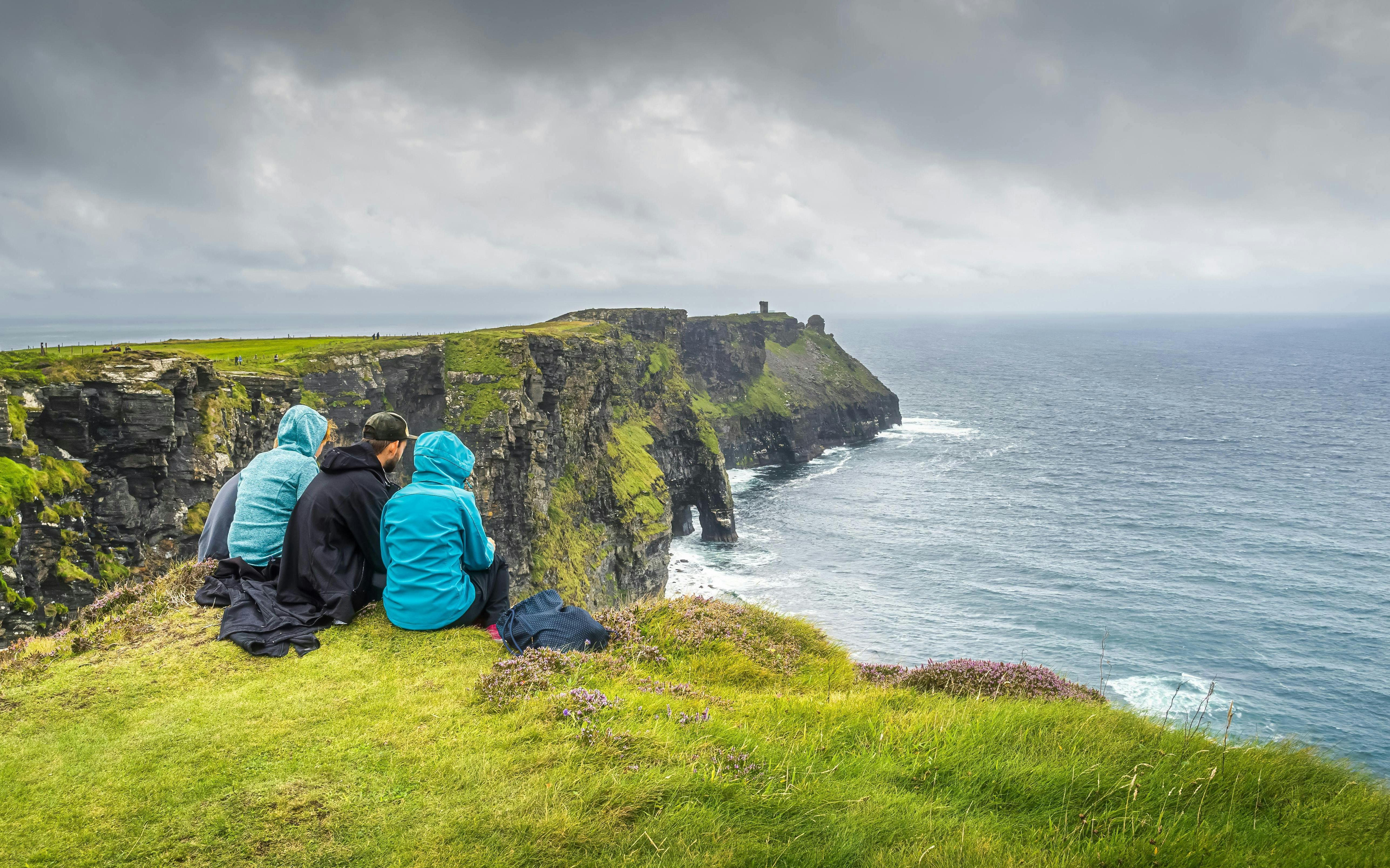 Visitors sitting on grassy edge overlooking Cliffs of Moher and Atlantic Ocean.