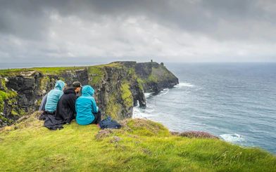 Visitors sitting on grassy edge overlooking Cliffs of Moher and Atlantic Ocean.