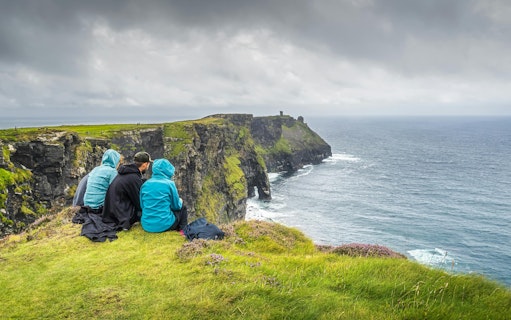 Visitors sitting on grassy edge overlooking Cliffs of Moher and Atlantic Ocean.