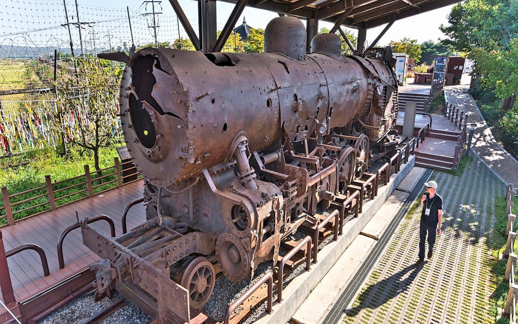 Rusty steam locomotive at DMZ, South Korea, with barbed wire fence and visitor nearby.