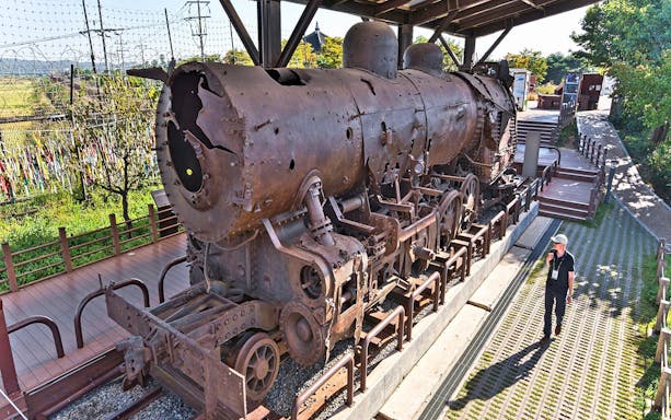 Rusty steam locomotive at DMZ, South Korea, with barbed wire fence and visitor nearby.