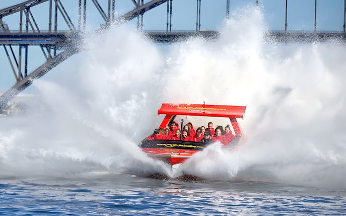 Jet boat splashing through water near Sydney Harbour Bridge with passengers.