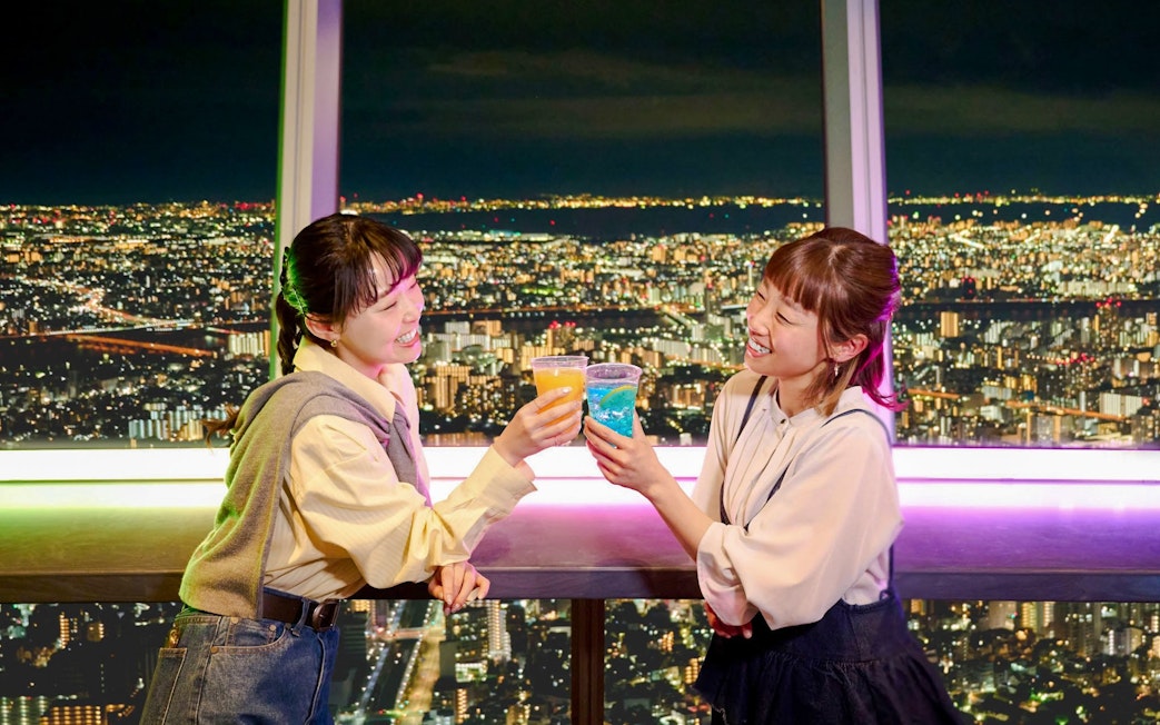Tourists toasting drinks at TOKYO SKYTREE Tembo Deck with city lights view.