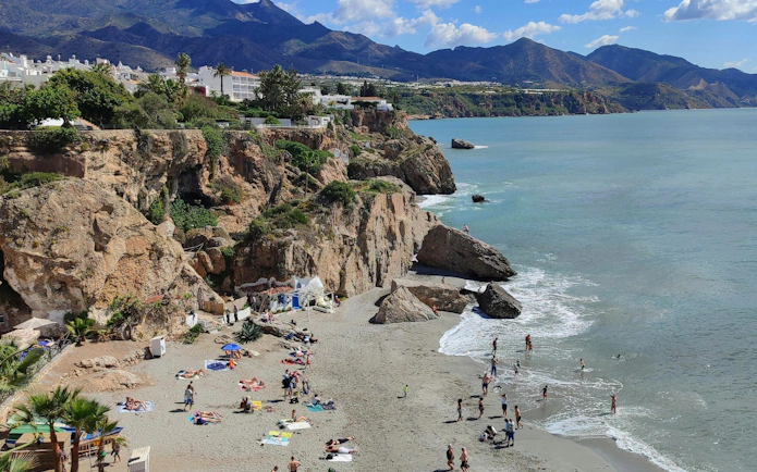Beachgoers enjoying the sun and sea near Balcón de Europa, Nerja, with rocky cliffs and mountains in the background.