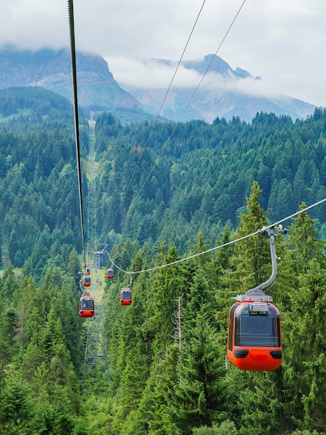 Pilatus Kulm cable cars ascending over lush green forest with mountains in the background.