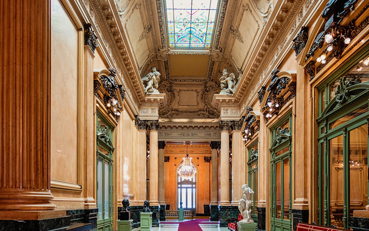 Teatro Colon interior with ornate columns, statues, and stained glass ceiling.