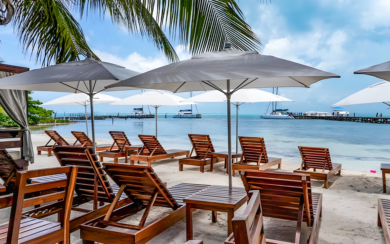 Beachfront lounge chairs under umbrellas with catamarans docked at Isla Mujeres.